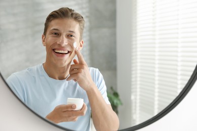Photo of Handsome young man applying cream onto his face near mirror at home