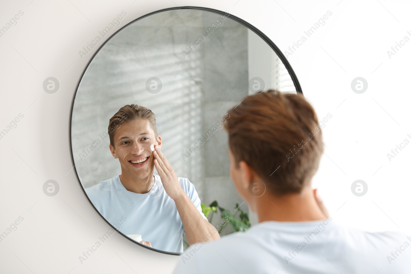 Photo of Handsome young man applying cream onto his face near mirror at home