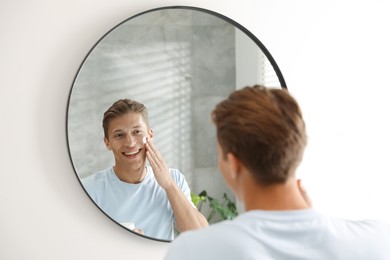 Photo of Handsome young man applying cream onto his face near mirror at home