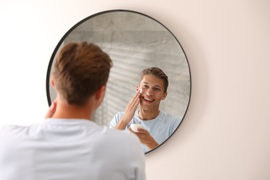 Photo of Handsome young man applying cream onto his face near mirror at home