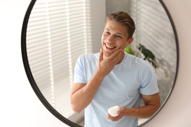 Photo of Handsome young man applying cream onto his face near mirror at home