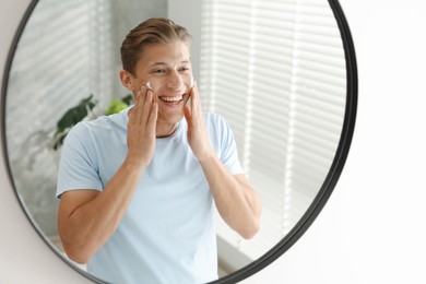 Photo of Handsome young man applying cream onto his face near mirror at home