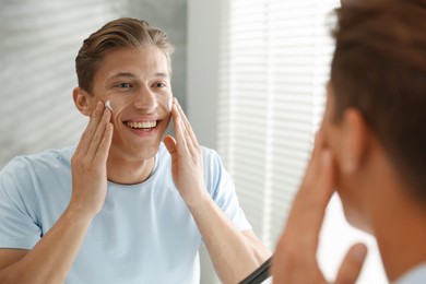 Photo of Handsome young man applying cream onto his face near mirror at home