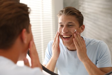 Photo of Handsome young man applying cream onto his face near mirror at home