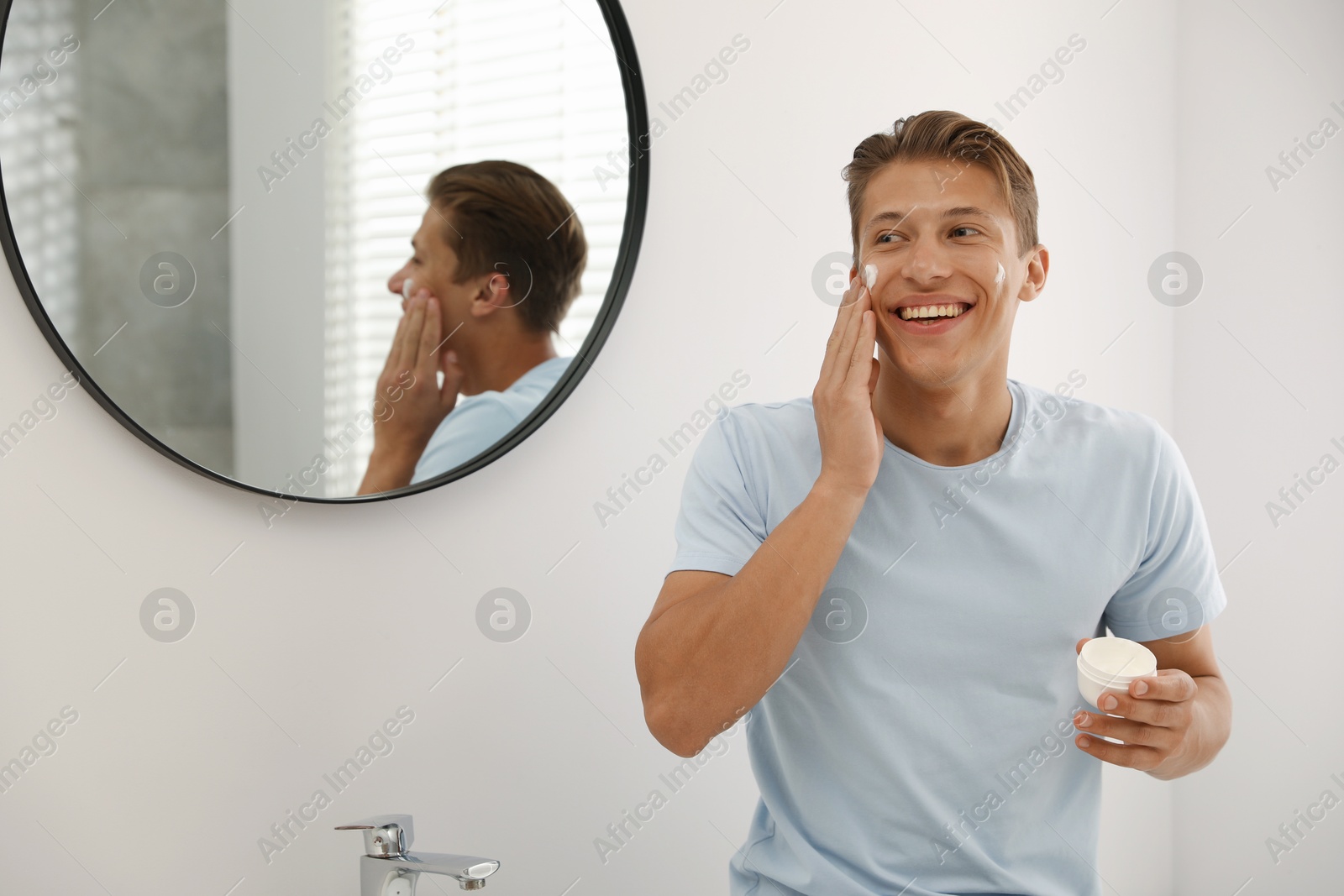 Photo of Handsome young man applying cream onto his face near mirror in bathroom