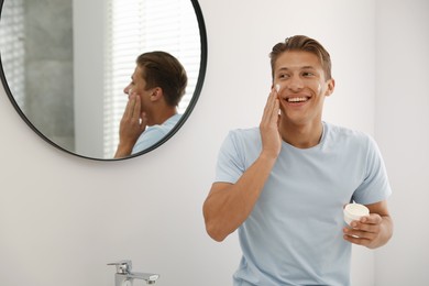 Photo of Handsome young man applying cream onto his face near mirror in bathroom