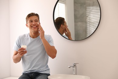 Photo of Handsome young man applying cream onto his face near mirror in bathroom