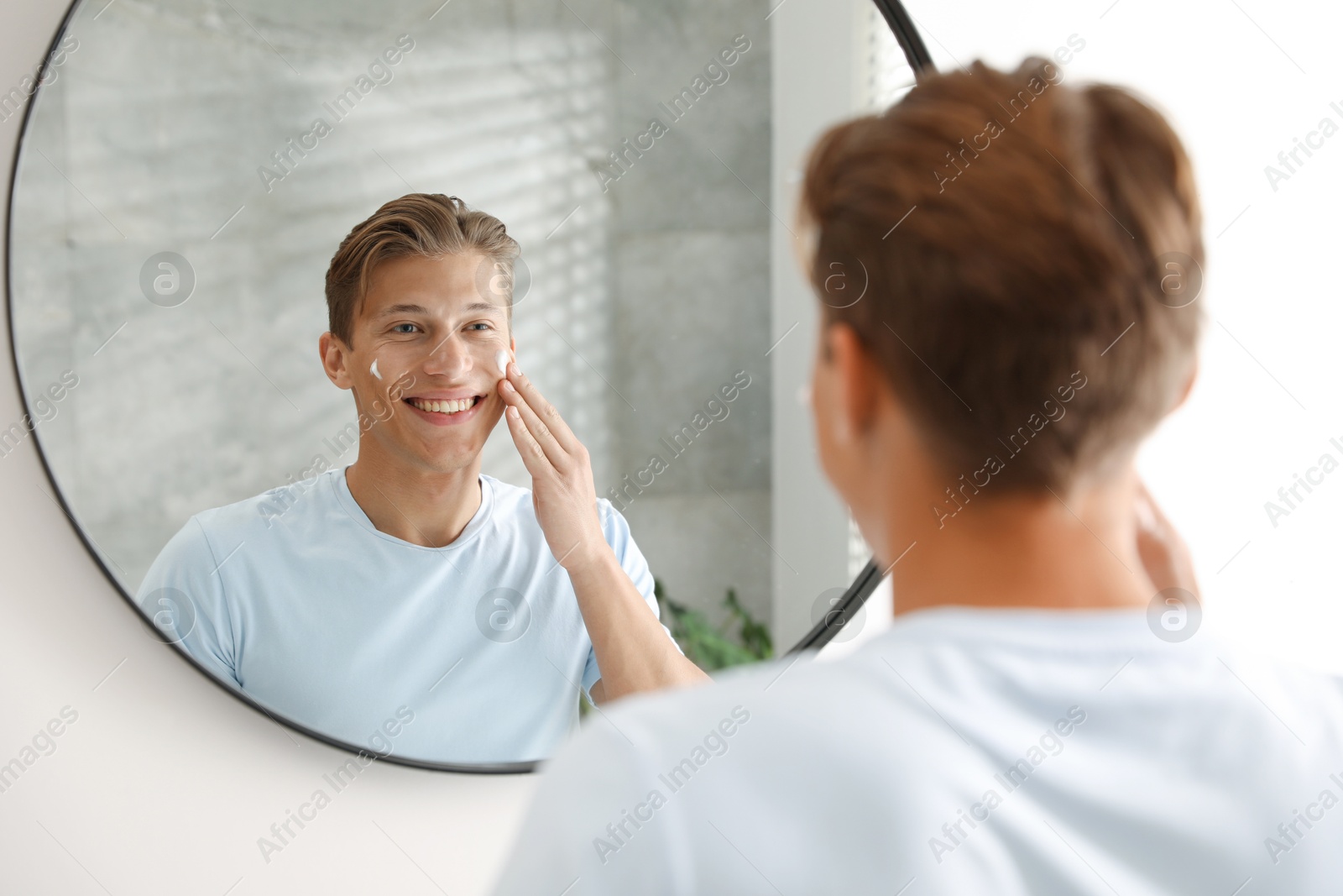 Photo of Handsome young man applying cream onto his face near mirror at home