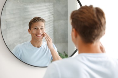 Photo of Handsome young man applying cream onto his face near mirror at home