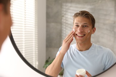 Photo of Handsome young man applying cream onto his face near mirror at home