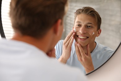 Photo of Handsome young man applying cream onto his face near mirror at home