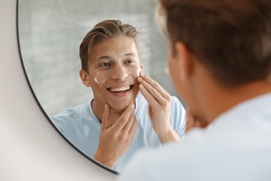 Photo of Handsome young man applying cream onto his face near mirror at home
