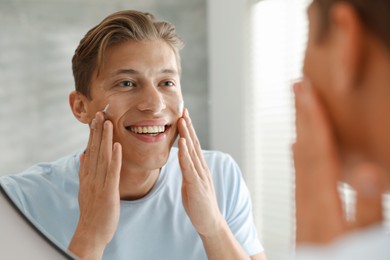 Photo of Handsome young man applying cream onto his face near mirror at home