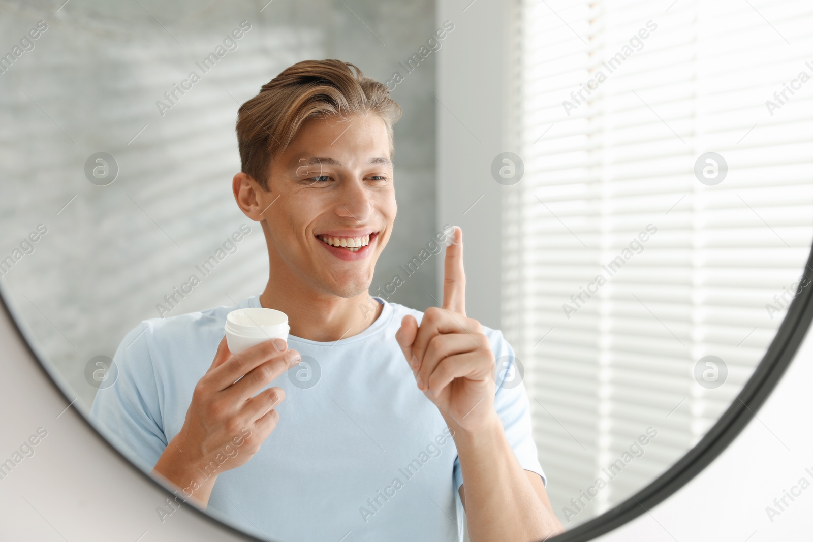 Photo of Handsome young man with jar of face cream near mirror at home