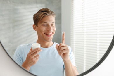 Photo of Handsome young man with jar of face cream near mirror at home