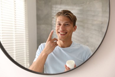 Photo of Handsome young man with jar of face cream near mirror at home