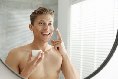 Photo of Handsome young man with jar of face cream near mirror at home
