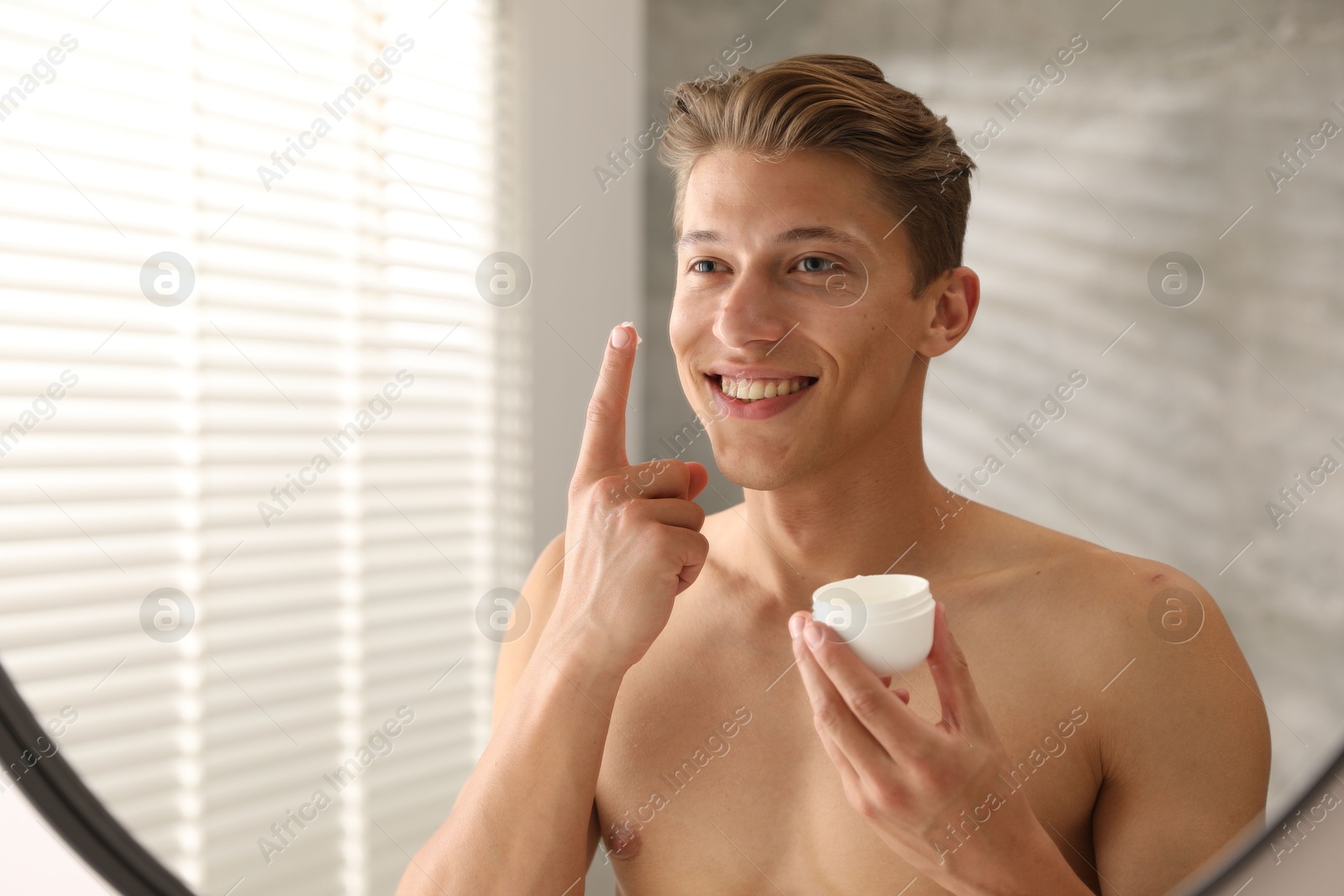 Photo of Handsome young man with jar of face cream near mirror at home