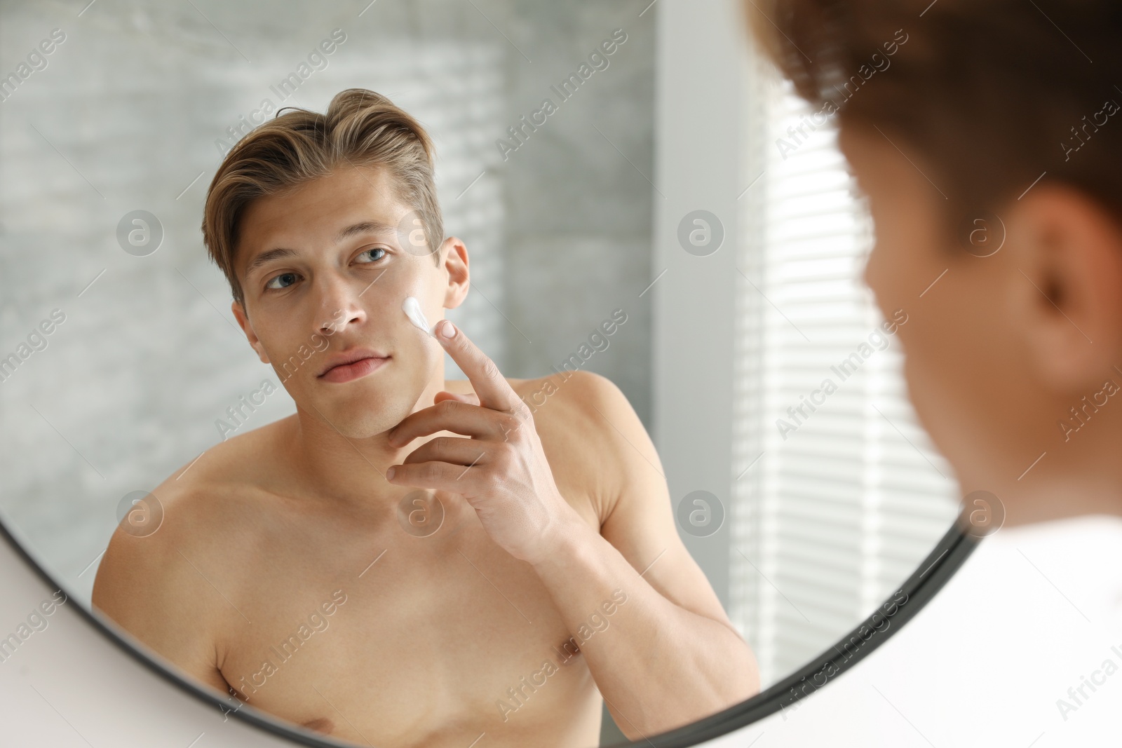 Photo of Handsome young man applying cream onto his face near mirror at home