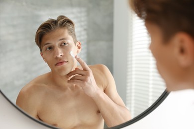 Photo of Handsome young man applying cream onto his face near mirror at home