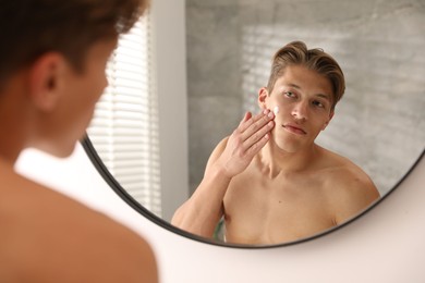 Photo of Handsome young man applying cream onto his face near mirror at home