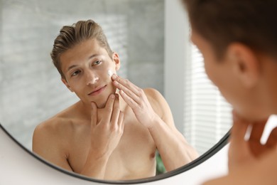 Photo of Handsome young man applying cream onto his face near mirror at home