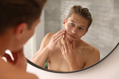 Photo of Handsome young man applying cream onto his face near mirror at home