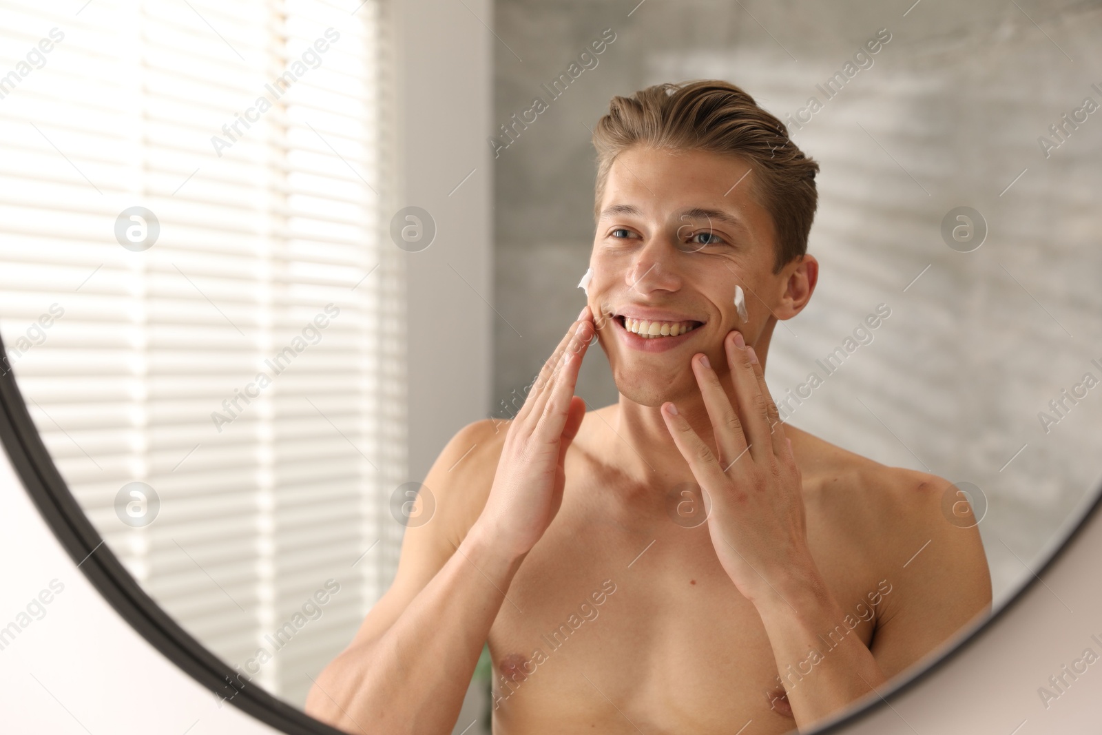 Photo of Handsome young man applying cream onto his face near mirror at home