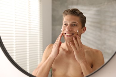 Photo of Handsome young man applying cream onto his face near mirror at home