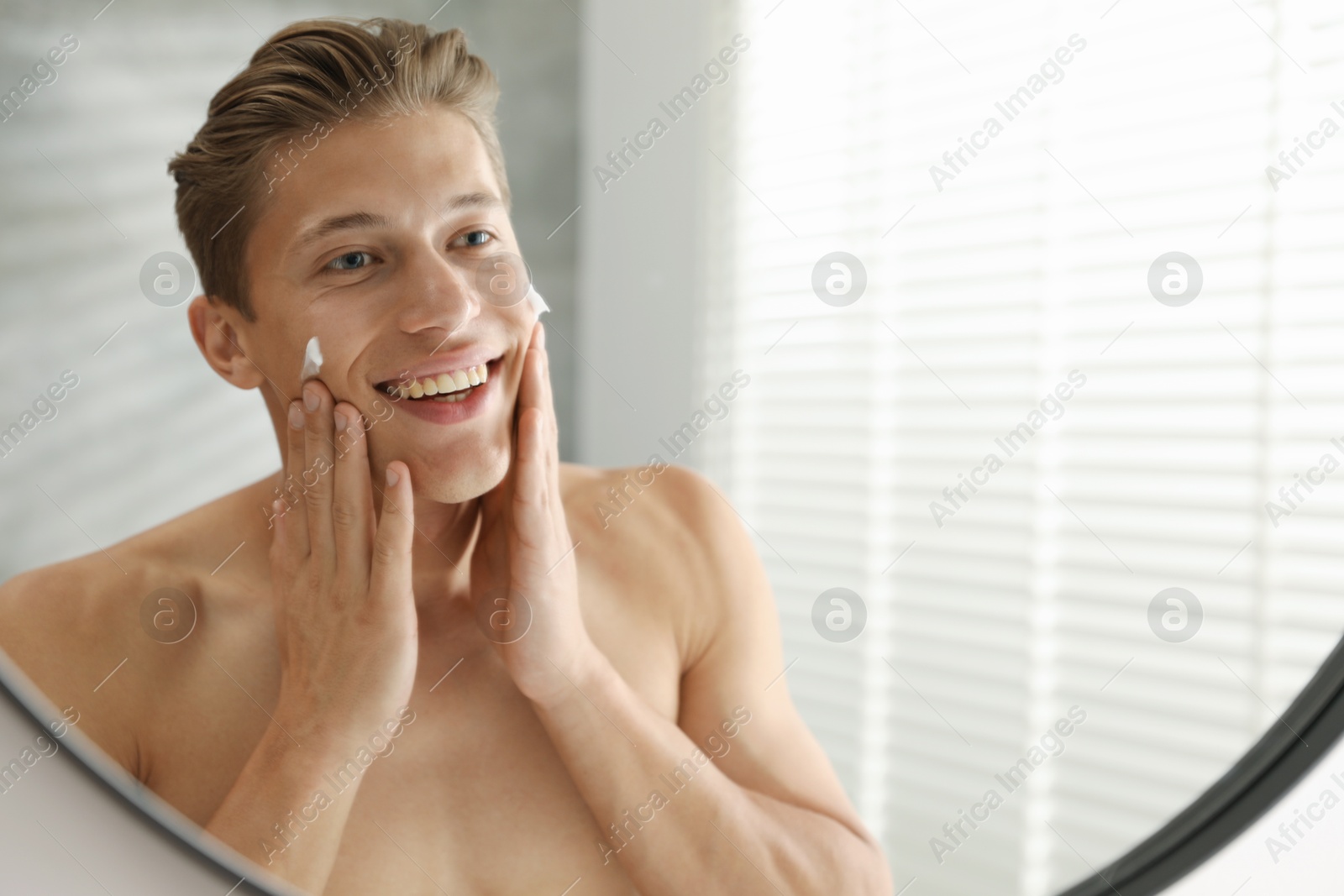 Photo of Handsome young man applying cream onto his face near mirror at home, space for text