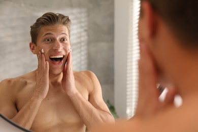 Photo of Handsome young man applying cream onto his face near mirror at home