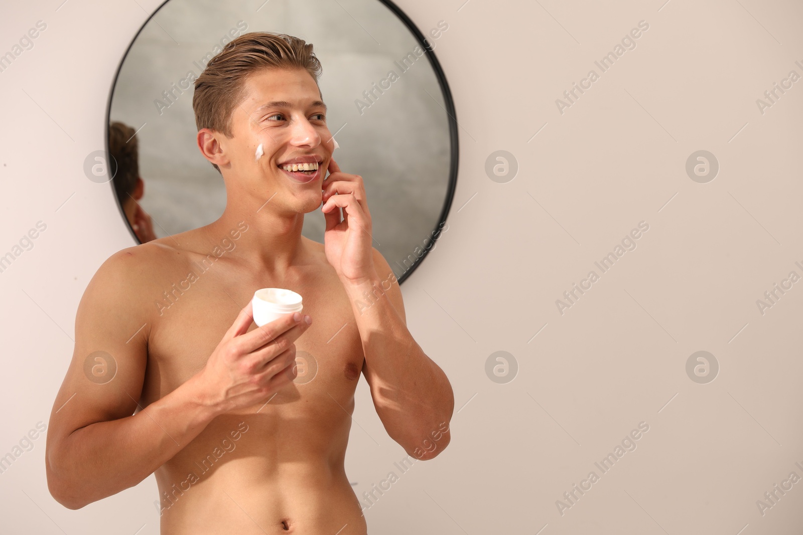 Photo of Handsome young man applying cream onto his face near mirror at home, space for text