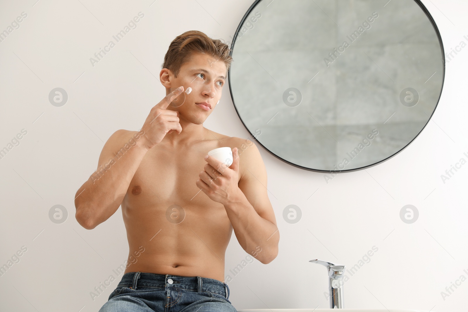 Photo of Handsome young man applying cream onto his face near mirror in bathroom