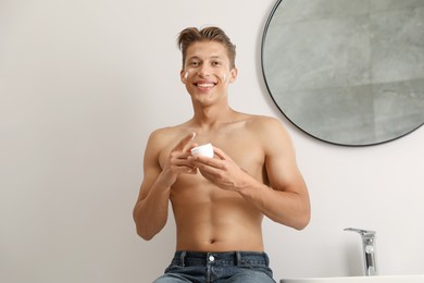 Photo of Handsome young man with jar of face cream near mirror in bathroom