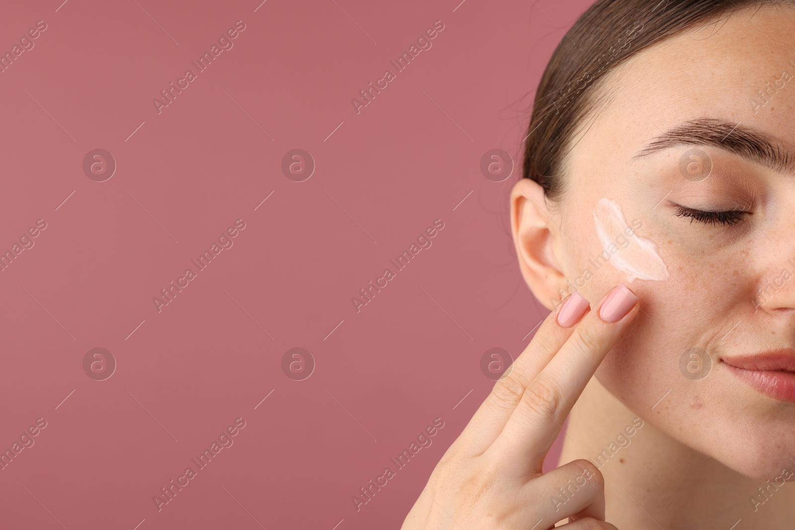 Photo of Beautiful woman applying cream onto her face on pink background, closeup. Space for text