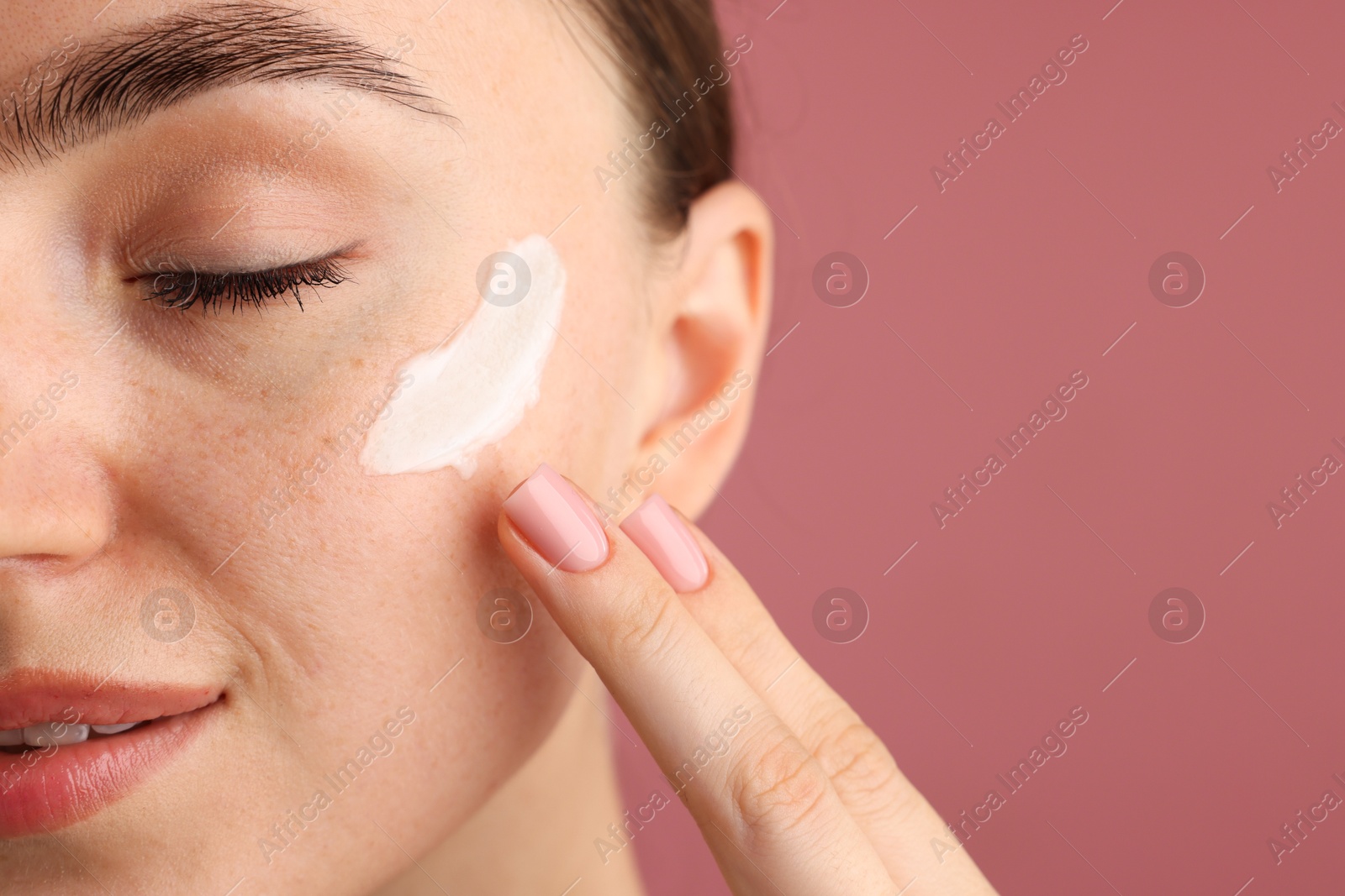 Photo of Beautiful woman applying cream onto her face on pink background, closeup. Space for text