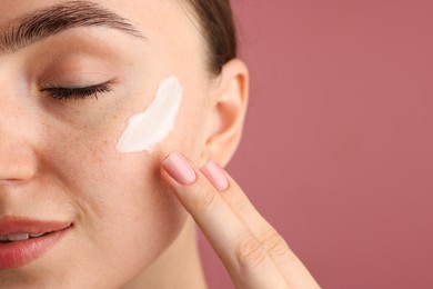 Photo of Beautiful woman applying cream onto her face on pink background, closeup. Space for text