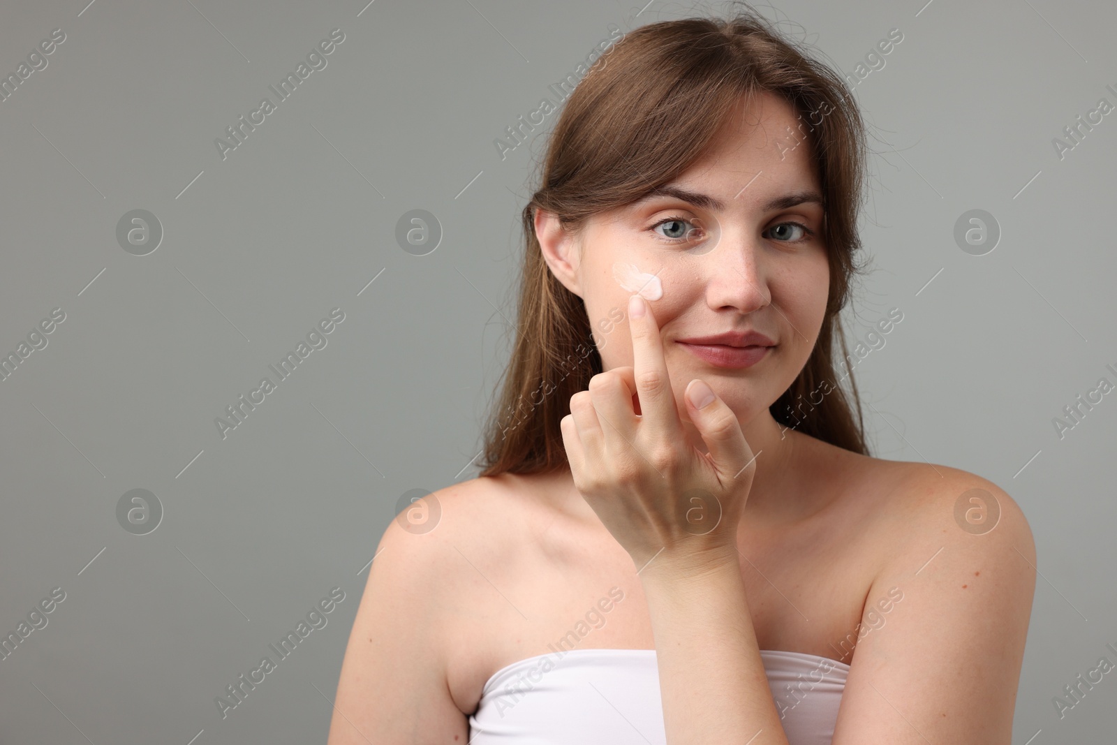 Photo of Beautiful woman applying cream onto her face on gray background. Space for text