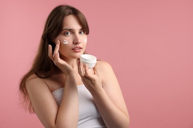 Photo of Beautiful woman applying cream onto her face on pink background. Space for text