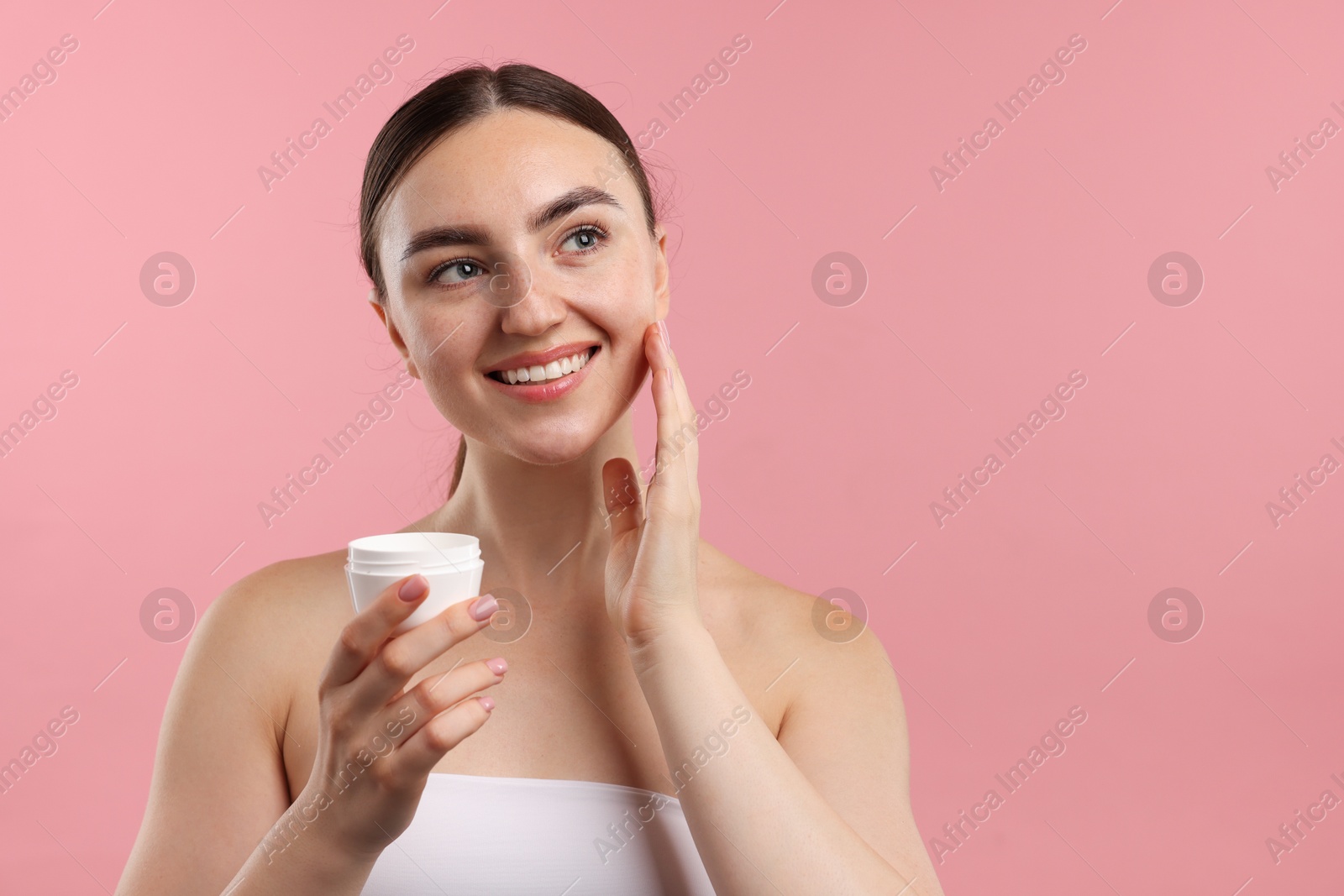 Photo of Beautiful woman applying cream onto her face on pink background. Space for text
