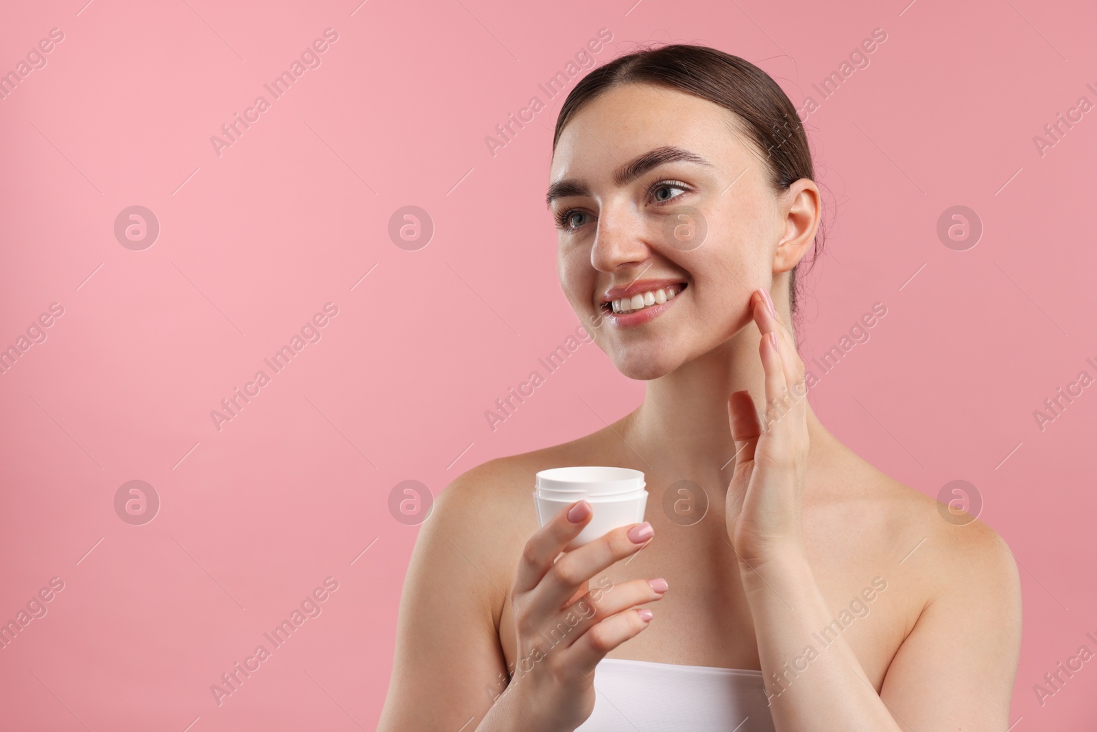 Photo of Beautiful woman applying cream onto her face on pink background. Space for text