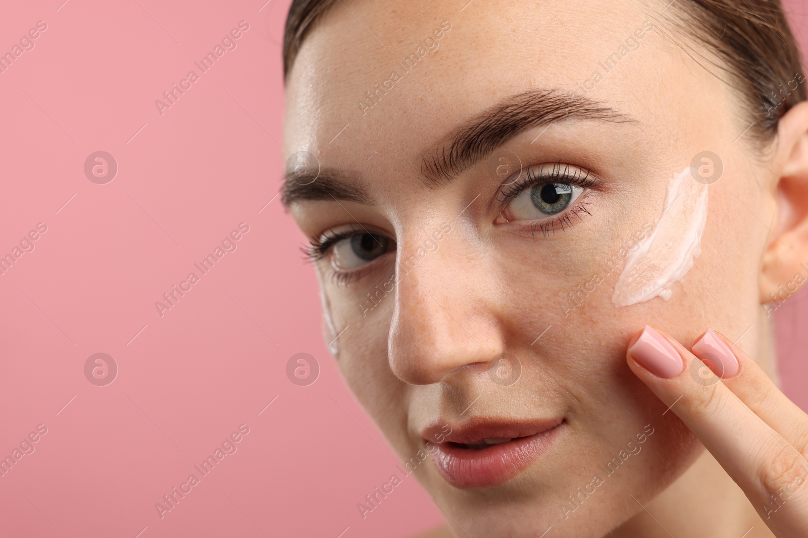 Photo of Beautiful woman with cream on her face against pink background, closeup. Space for text