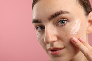Photo of Beautiful woman with cream on her face against pink background, closeup. Space for text