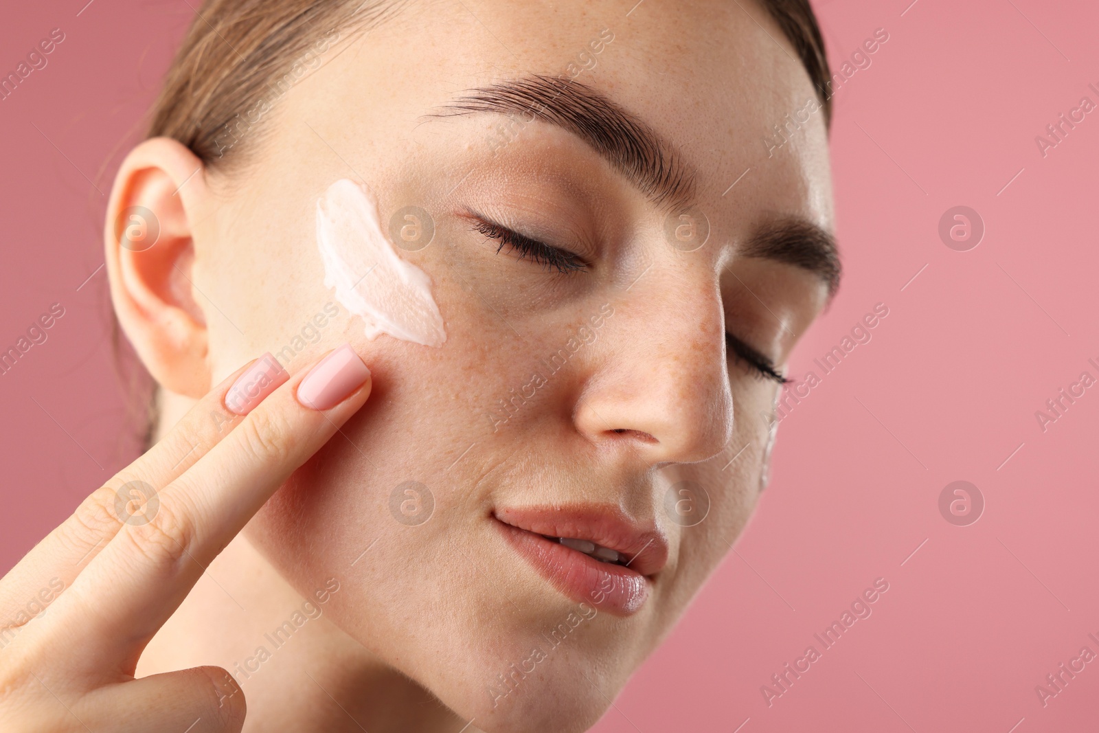 Photo of Beautiful woman applying cream onto her face on pink background, closeup