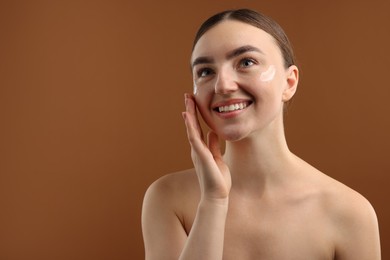 Photo of Beautiful woman applying cream onto her face on brown background. Space for text