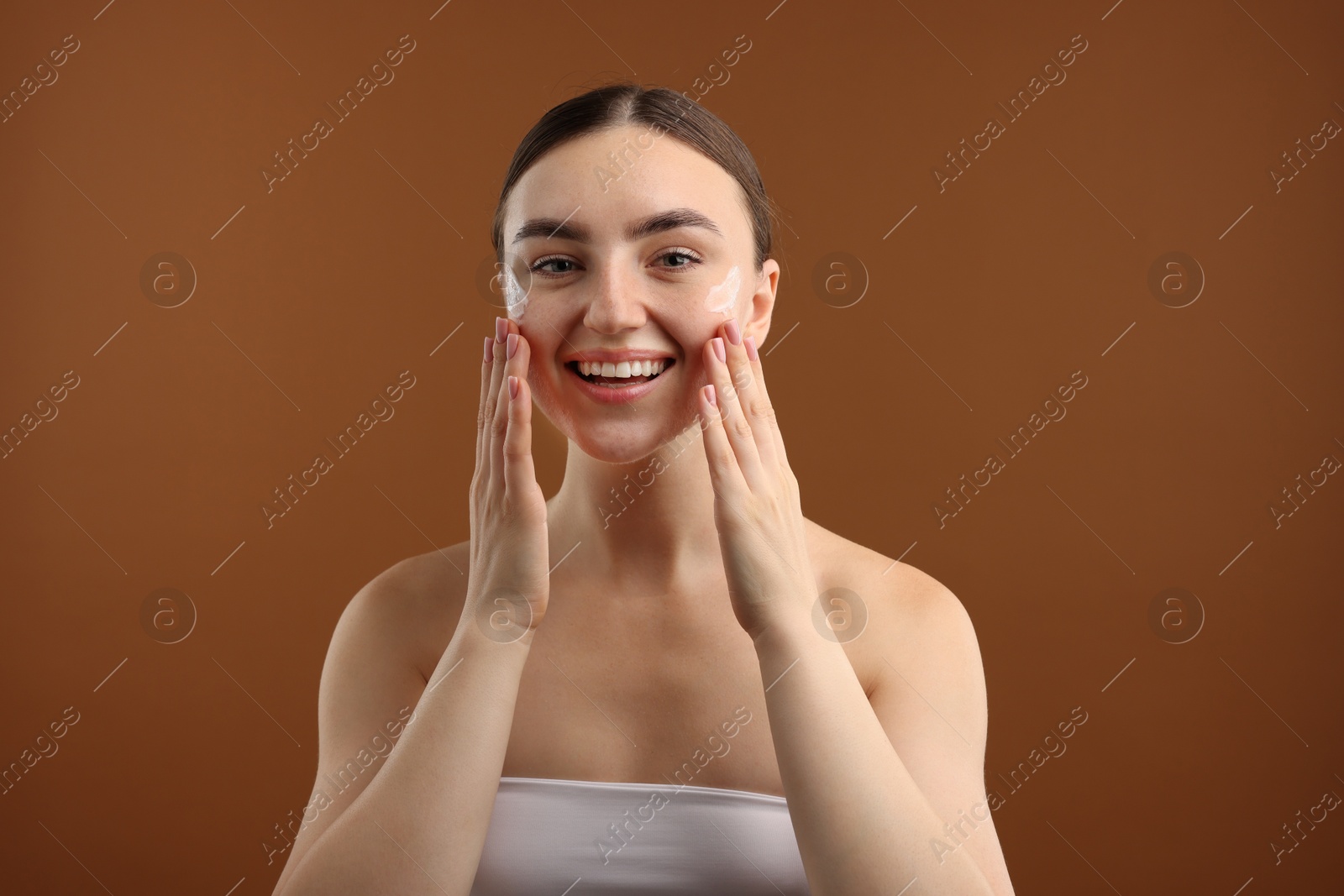 Photo of Beautiful woman applying cream onto her face on brown background