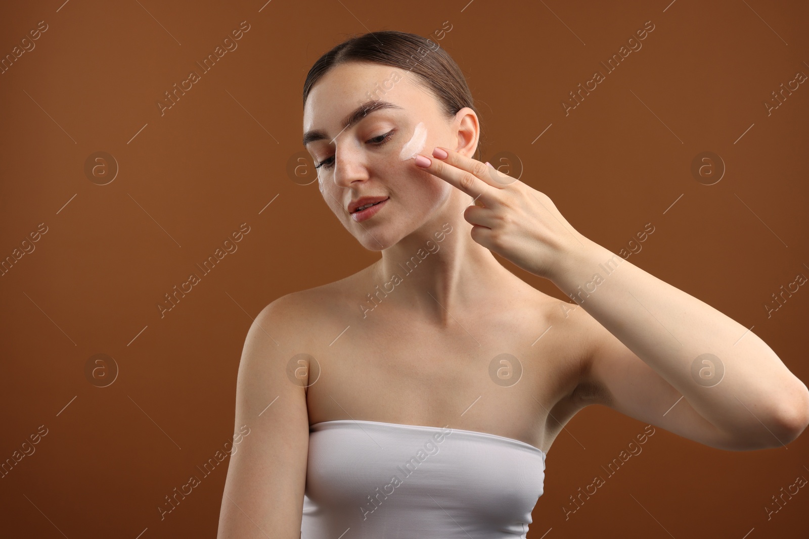 Photo of Beautiful woman applying cream onto her face on brown background