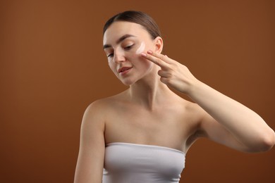 Photo of Beautiful woman applying cream onto her face on brown background