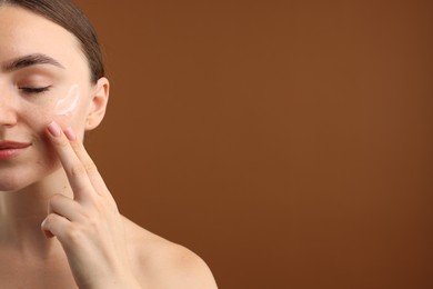 Photo of Beautiful woman applying cream onto her face on brown background, closeup. Space for text