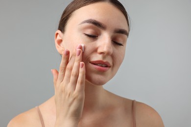 Photo of Beautiful woman applying cream onto her face on gray background, closeup